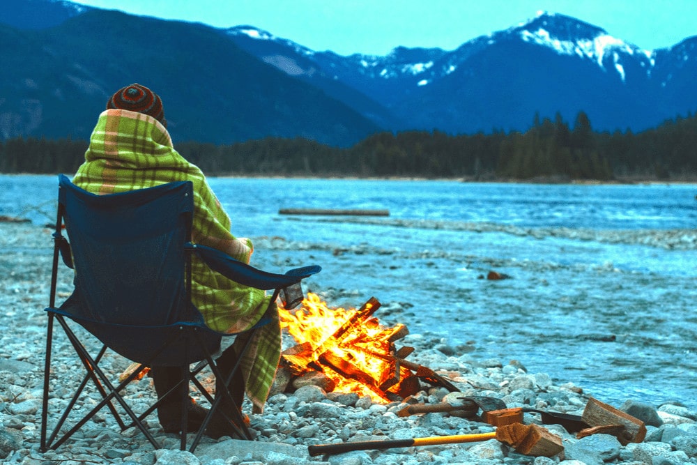 A person sitting by the campfire covered in  a camping blanket 