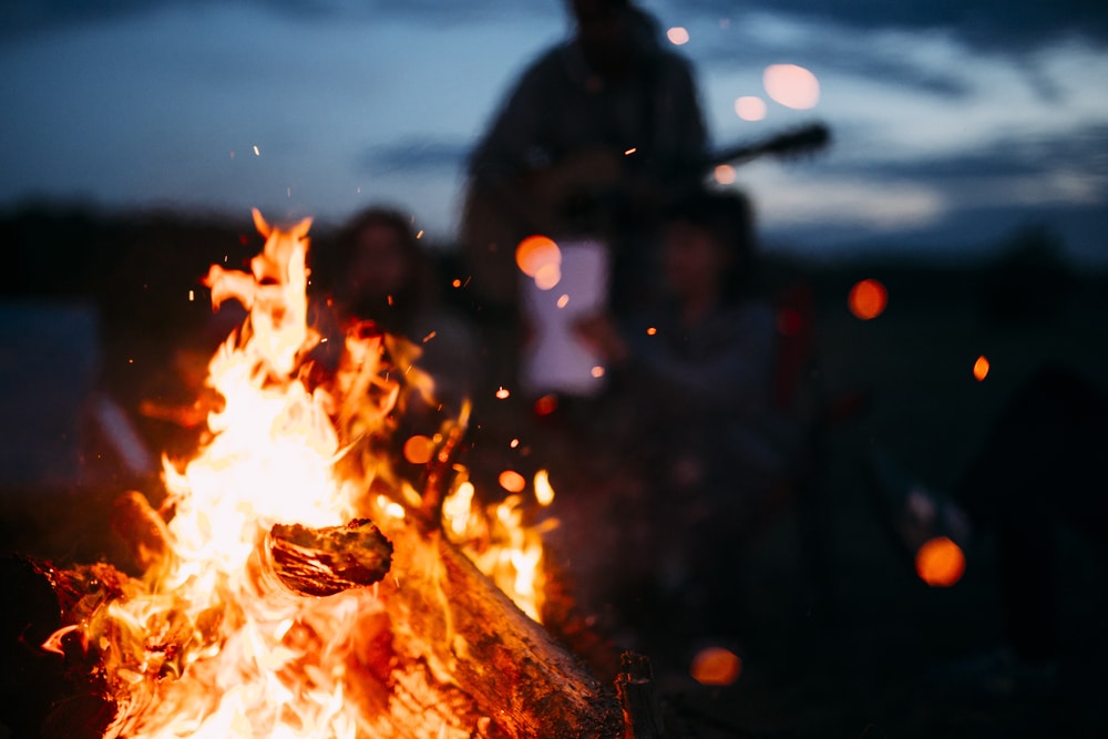close up image of a campfire and spark flying around with group of friends at the background