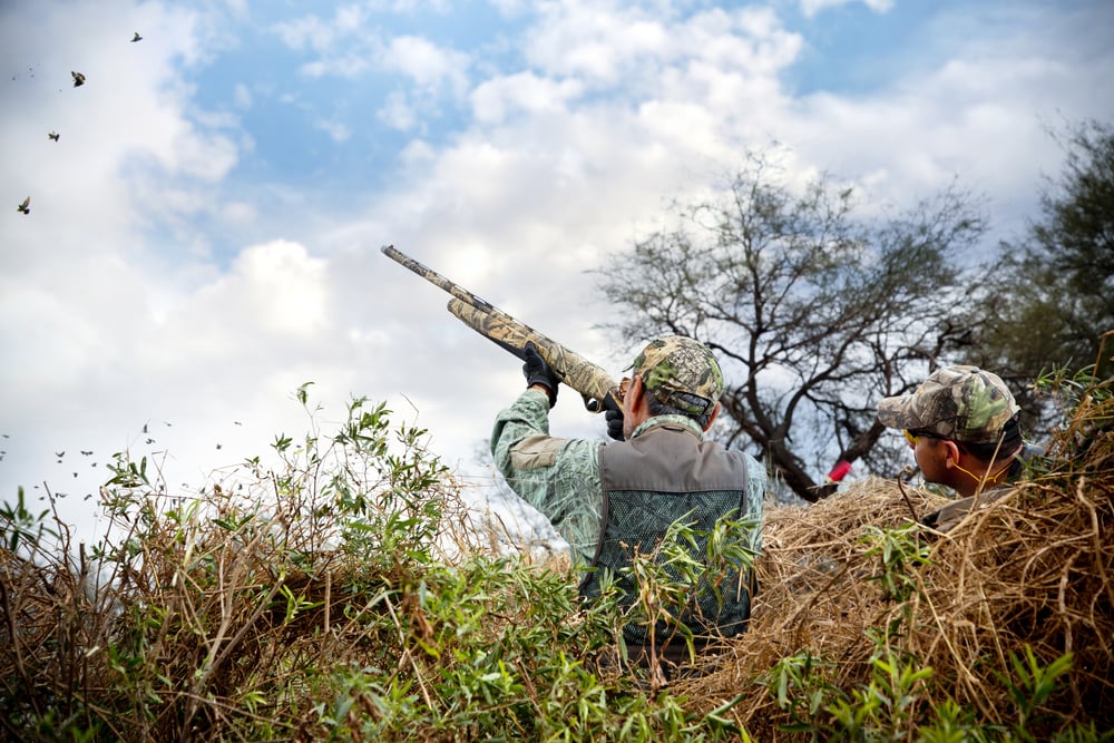 two men hiding in grass while hunting birds in the sky