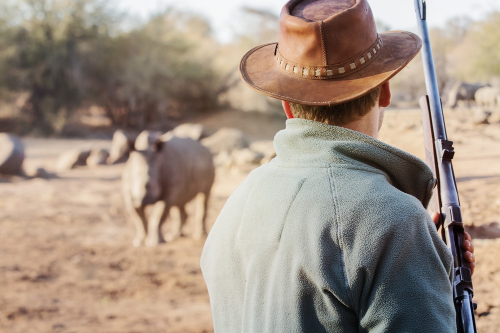 a man holding a firearm near rhinos in a savanna