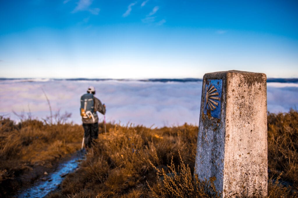 Pilgrim walking in Camino de Santiago, over a sea of clouds and a stone with the shell symbol